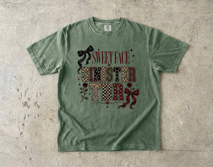 A green graphic t-shirt laid flat on a textured concrete surface. The shirt features a decorative graphic on the front with the words 'SWEET FACE SINISTER' in a patchwork pattern, accented with dark red bows and small floral details.
