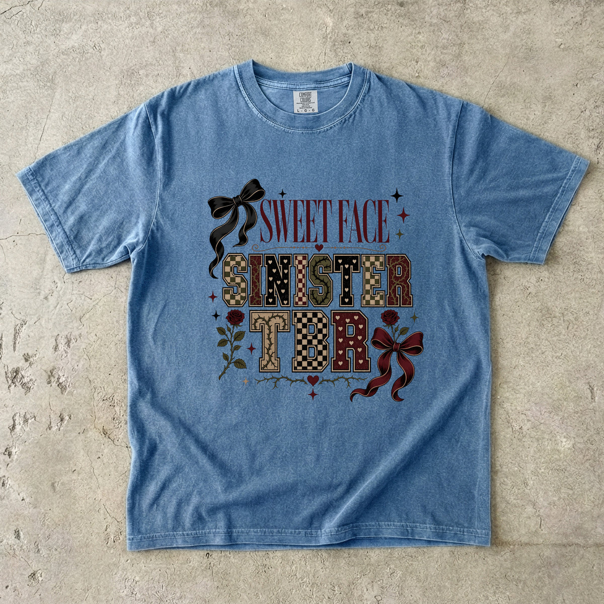 A light blue short-sleeve graphic t-shirt laid flat on a textured concrete surface. The graphic features the words 'SWEET FACE SINISTER' in a mix of red, brown, and patterned fonts, decorated with black and red bows, roses, and small stars.