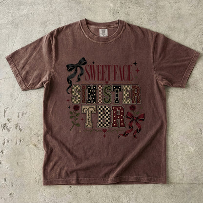 A washed brown short-sleeve t-shirt with a gothic-style graphic reading 'SWEET FACE SINISTER TONIGHT' in patterned letters, decorated with bows and roses, laid flat on a textured concrete surface.