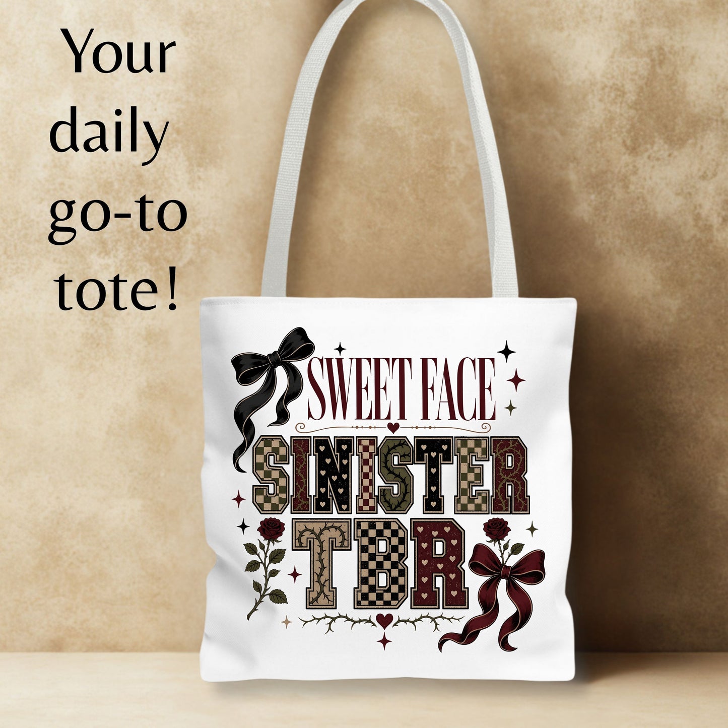 A white canvas tote bag with white shoulder straps, featuring a decorative printed design that reads 'SWEET FACE SINISTER TBR' with bows, roses, and patterned lettering. The bag is shown against a textured beige background with promotional text on the left reading 'Your daily go-to tote!'.