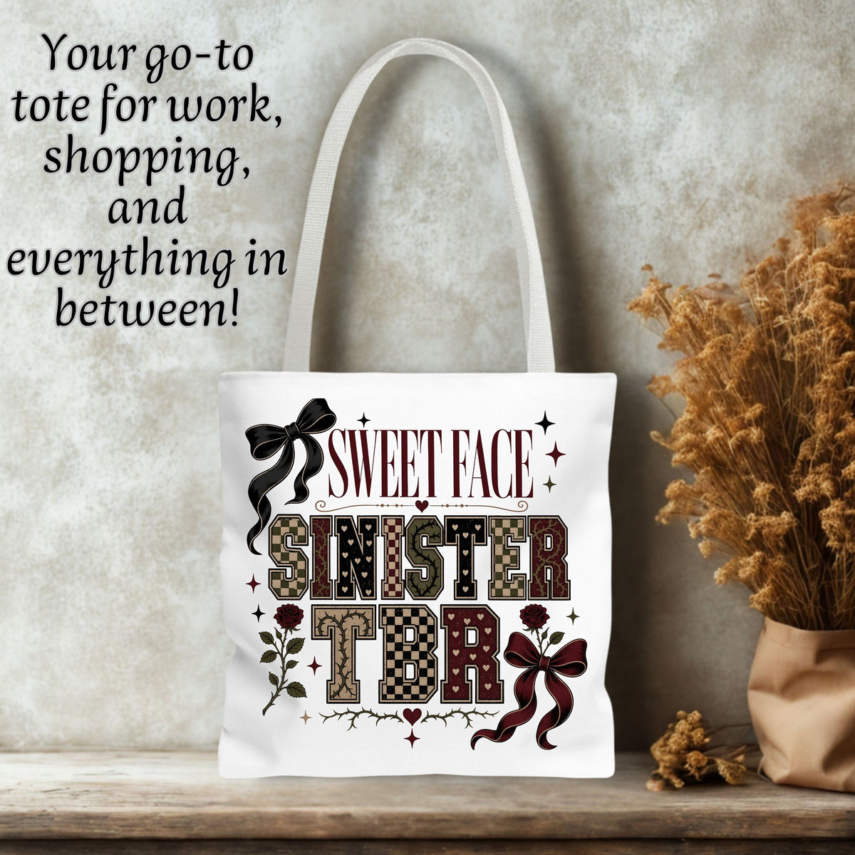 A white canvas tote bag with a gothic-style 'Sweet Face Sinister' graphic, displayed on a wooden shelf next to dried flowers against a textured gray wall. Black text in the upper left reads 'Your go-to tote for work, shopping, and everything in between!'.