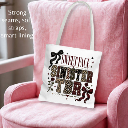 A white canvas tote bag with a 'Sweet Sister Face' graphic print, displayed on a pink armchair. The bag has white shoulder straps and features a graphic with the words 'SWEET FACE SINISTER TONIGHT' in patterned and black lettering, accented with bows and stars.