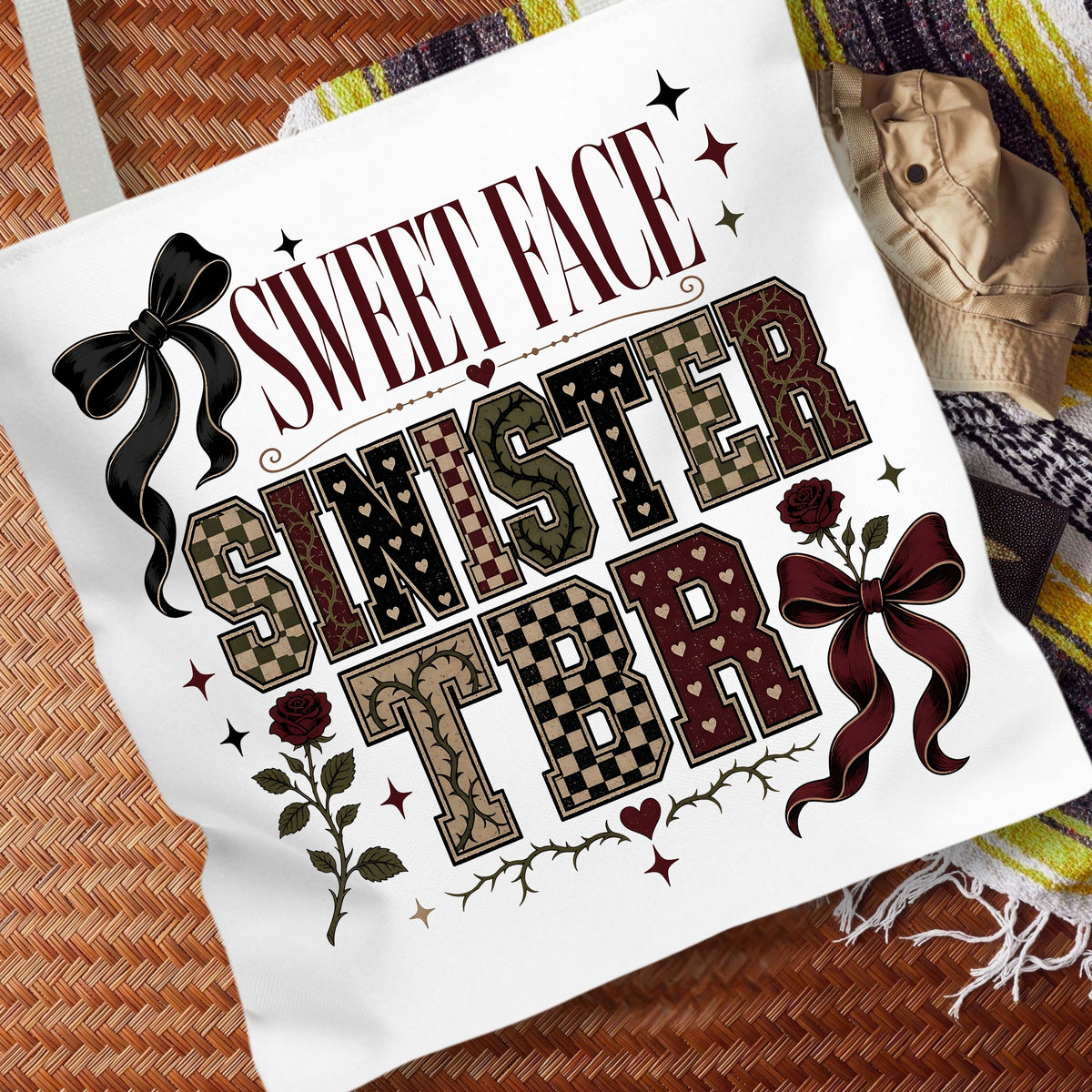 A white canvas tote bag featuring a gothic-style graphic print that reads 'SWEET FACE SINISTER TBR' with decorative bows, roses, stars, and patterned lettering, styled on a woven brown surface next to a beige hat and a striped blanket.