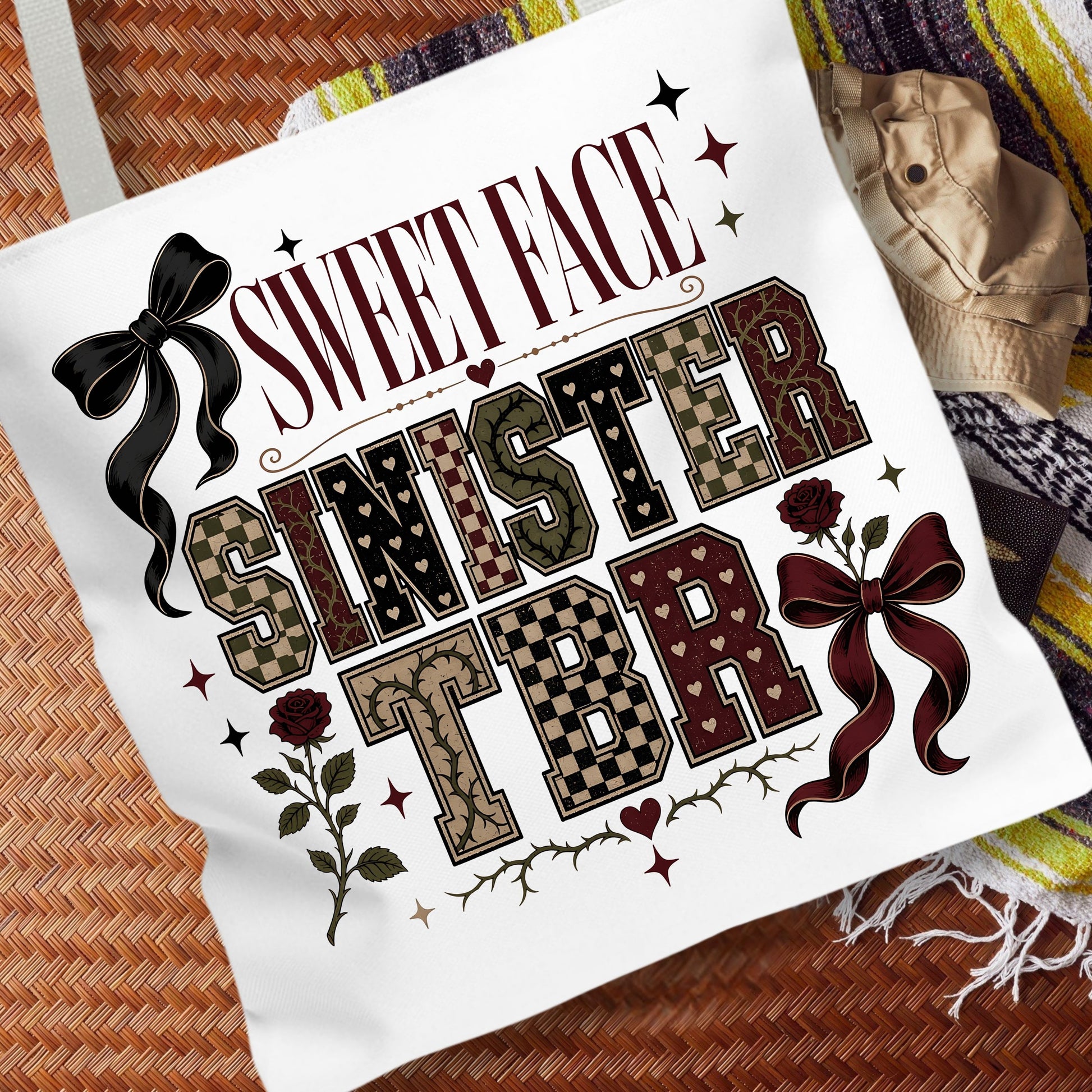 A white canvas tote bag featuring a gothic-style graphic print that reads 'SWEET FACE SINISTER TBR' with decorative bows, roses, stars, and patterned lettering, styled on a woven brown surface next to a beige hat and a striped blanket.