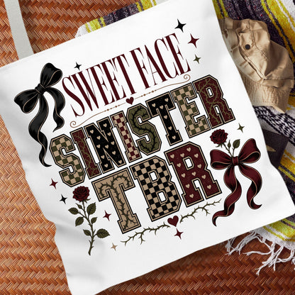 A white canvas tote bag featuring a gothic-style graphic print that reads 'SWEET FACE SINISTER TBR' with decorative bows, roses, stars, and patterned lettering, styled on a woven brown surface next to a beige hat and a striped blanket.