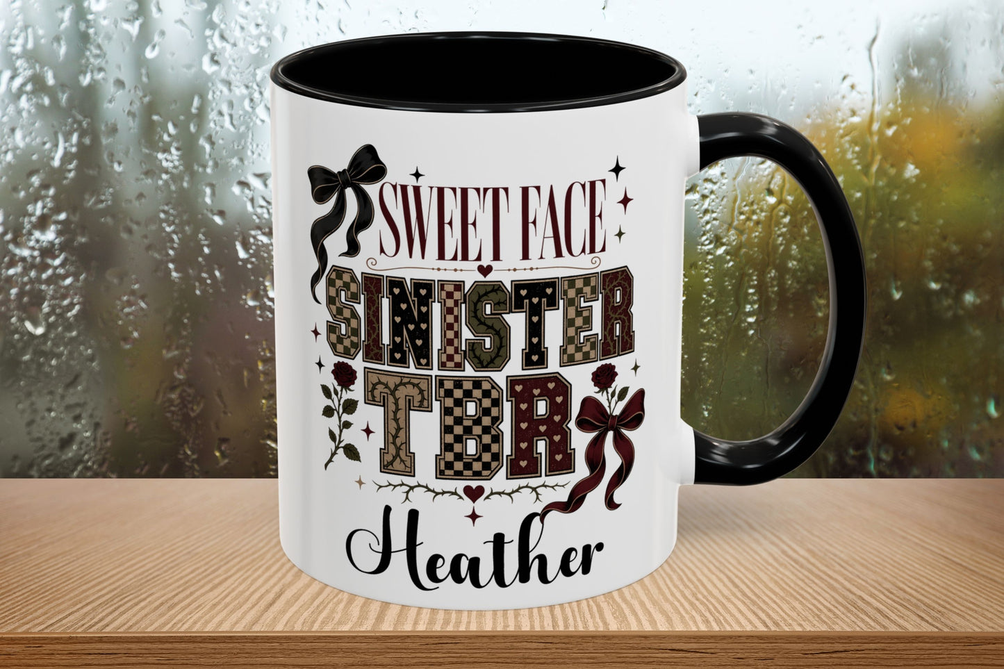 A white ceramic mug with a black handle and interior, featuring a custom printed design with the text 'SWEET FACE SINISTER TDR' and 'Heather' in black and patterned lettering. The mug is shown on a wooden surface with a blurred, rainy window background.