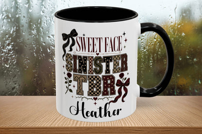 A white ceramic mug with a black handle and interior, featuring a custom printed design with the text 'SWEET FACE SINISTER TDR' and 'Heather' in black and patterned lettering. The mug is shown on a wooden surface with a blurred, rainy window background.