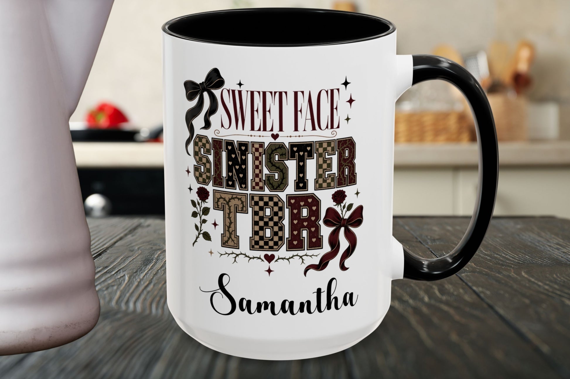 A white ceramic mug with a black handle and black interior, featuring a custom holiday-themed graphic that reads 'SWEET FACE SINISTER TDR' with patterned letters and bows, and the name 'Samantha' at the bottom. The mug is shown on a dark wooden table with a blurred kitchen background.
