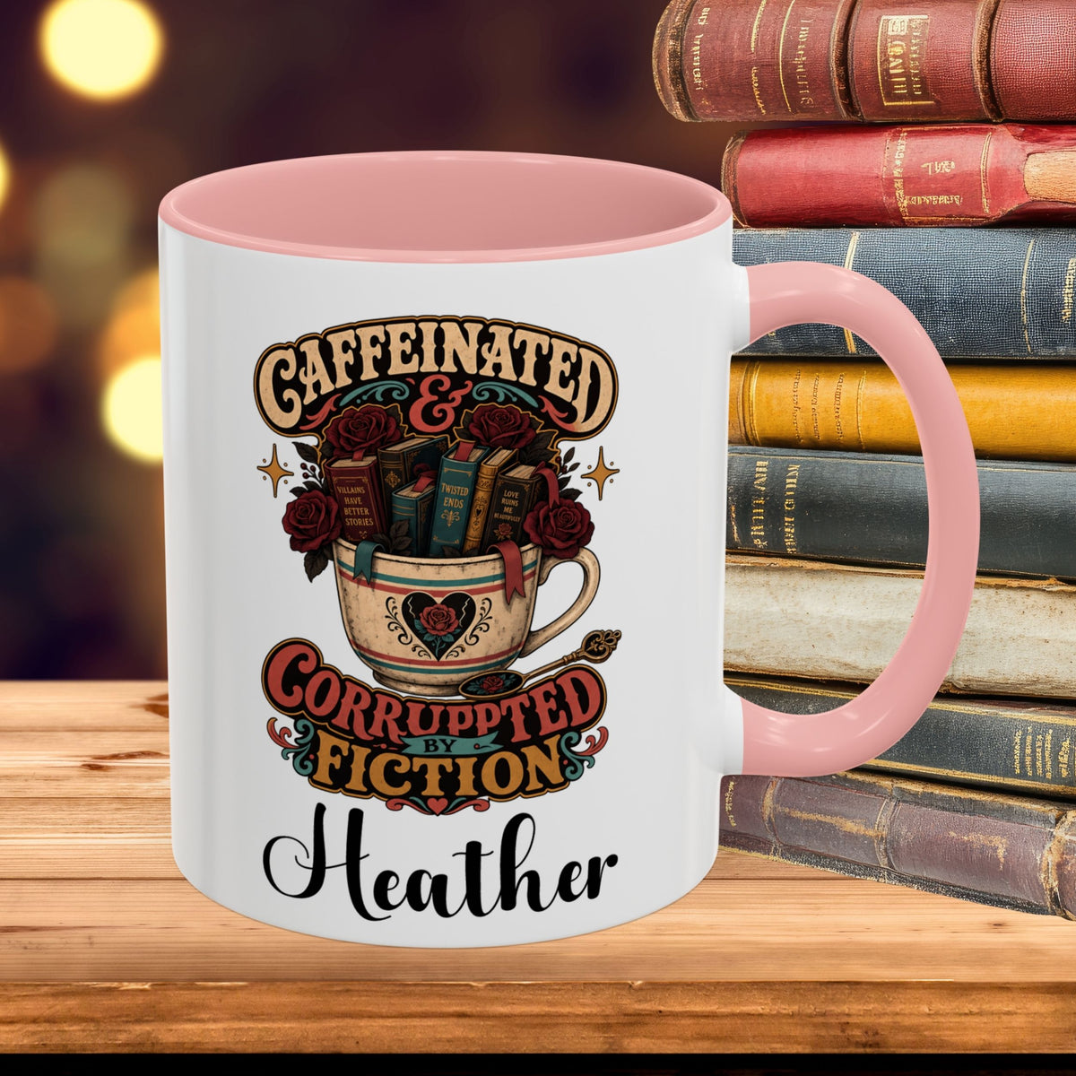 A white ceramic mug with a pink handle and interior, featuring a vintage-style graphic of a teacup filled with books and roses and the text 'CAFFEINATED & CORRUPTED FICTION Heather'. The mug is placed on a wooden surface with a stack of old books and a blurred warm background.