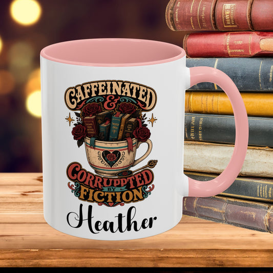 A white ceramic mug with a pink handle and interior, featuring a vintage-style graphic of a teacup filled with books and roses and the text 'CAFFEINATED & CORRUPTED FICTION Heather'. The mug is placed on a wooden surface with a stack of old books and a blurred warm background.