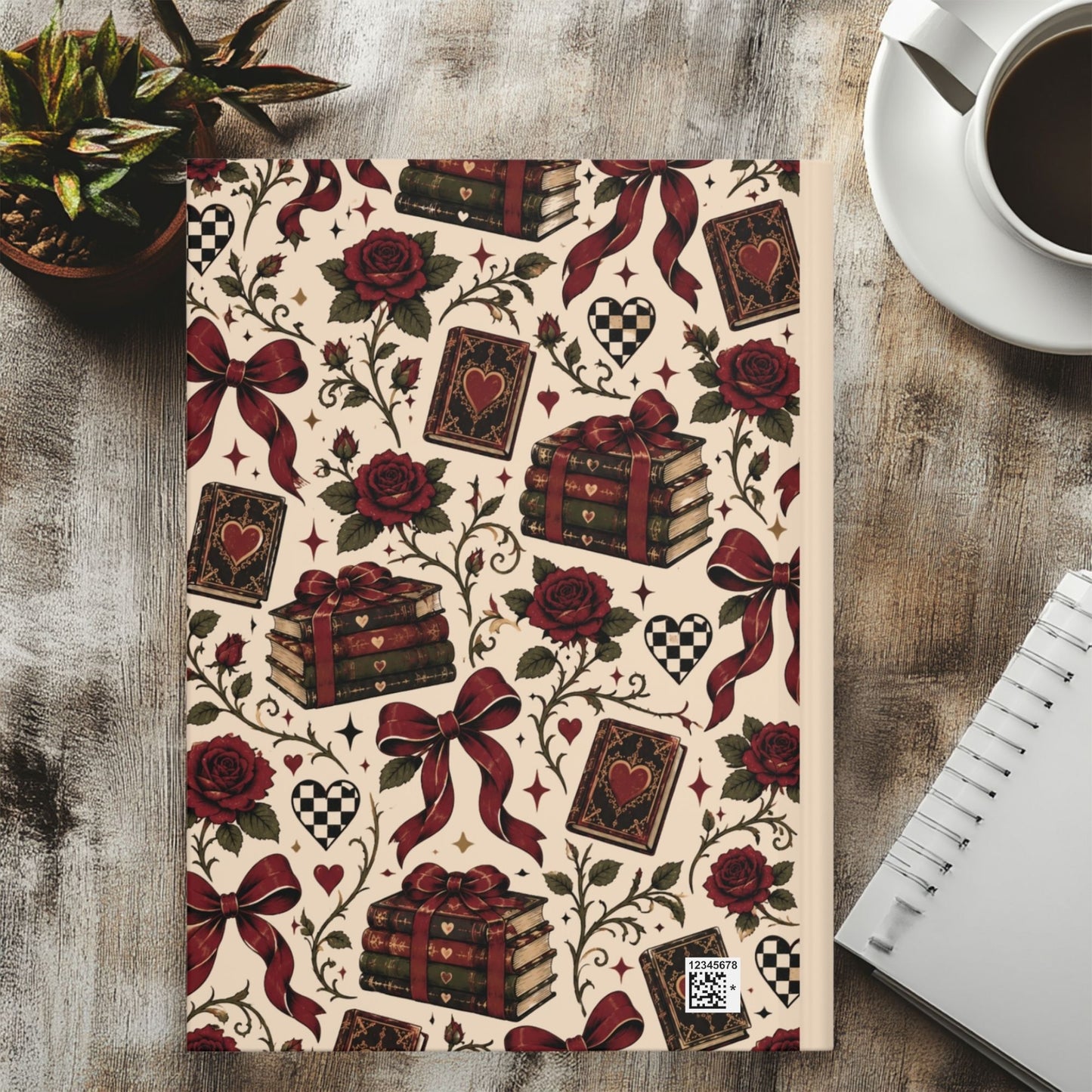 A notebook with a vintage-style pattern featuring red roses, playing cards, stacked books tied with red ribbons, hearts, and checkered motifs on a cream background, styled on a wooden tabletop with a coffee cup, succulent, and spiral notebook nearby.