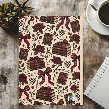 A notebook with a vintage-style pattern featuring red roses, playing cards, stacked books tied with red ribbons, hearts, and checkered motifs on a cream background, styled on a wooden tabletop with a coffee cup, succulent, and spiral notebook nearby.