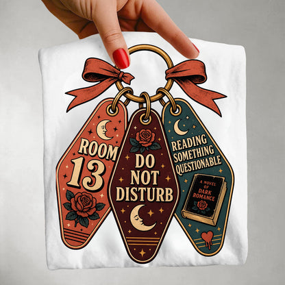 A folded white T-shirt is held by a hand with red nail polish. The shirt features a graphic print of three keychains with gothic-themed designs: one orange keychain says 'ROOM 13' with a moon and rose, one dark brown keychain says 'DO NOT DISTURB' with a rose and moon, and one teal keychain says 'READING SOMETHING QUESTIONABLE' with a book and moon.