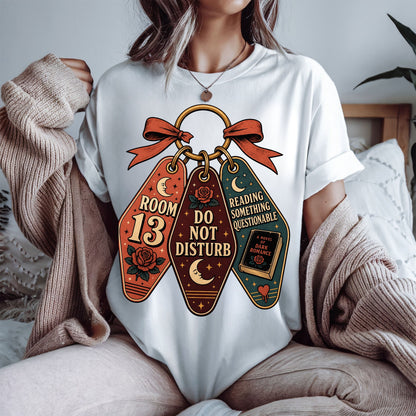 A woman wearing a white graphic t-shirt with a vintage-style keychain design featuring three charms that read 'ROOM 13', 'DO NOT DISTURB', and 'READING SOMETHING QUESTIONABLE'. She has a beige knit sweater draped off her shoulders in a cozy indoor setting.