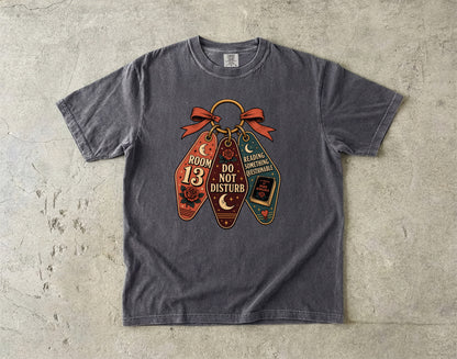 A dark gray heather t-shirt laid flat on a textured concrete surface, featuring a graphic of three hanging coffin-shaped tags with Halloween-themed text and illustrations.