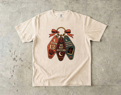 A cream-colored short-sleeve t-shirt laid flat on a textured gray concrete surface. The front of the shirt features a graphic print of three vintage-style book keychains tied together with a red ribbon.