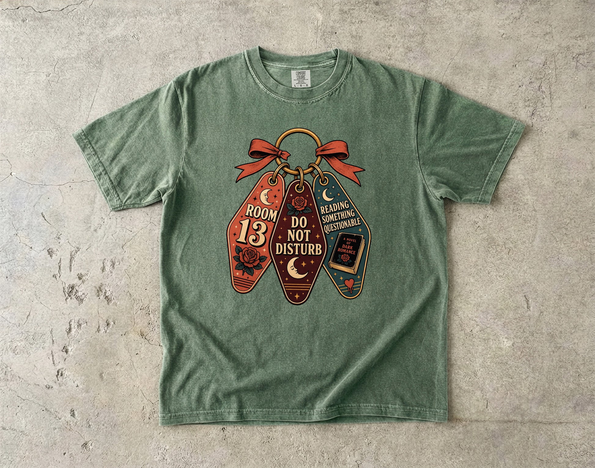 A sage green short-sleeve t-shirt with a graphic print of three Halloween-themed keychains on a textured concrete background. The keychains feature a skull, a book with a crescent moon, and a book with a witch hat, with text reading 'ROOM 13', 'DO NOT DISTURB', and 'READING COMFORTABLE FOREVER'.