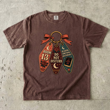 A brown short-sleeve t-shirt laid flat on a textured gray concrete surface. The shirt features a graphic print of three coffin-shaped keychains tied together with a red ribbon.