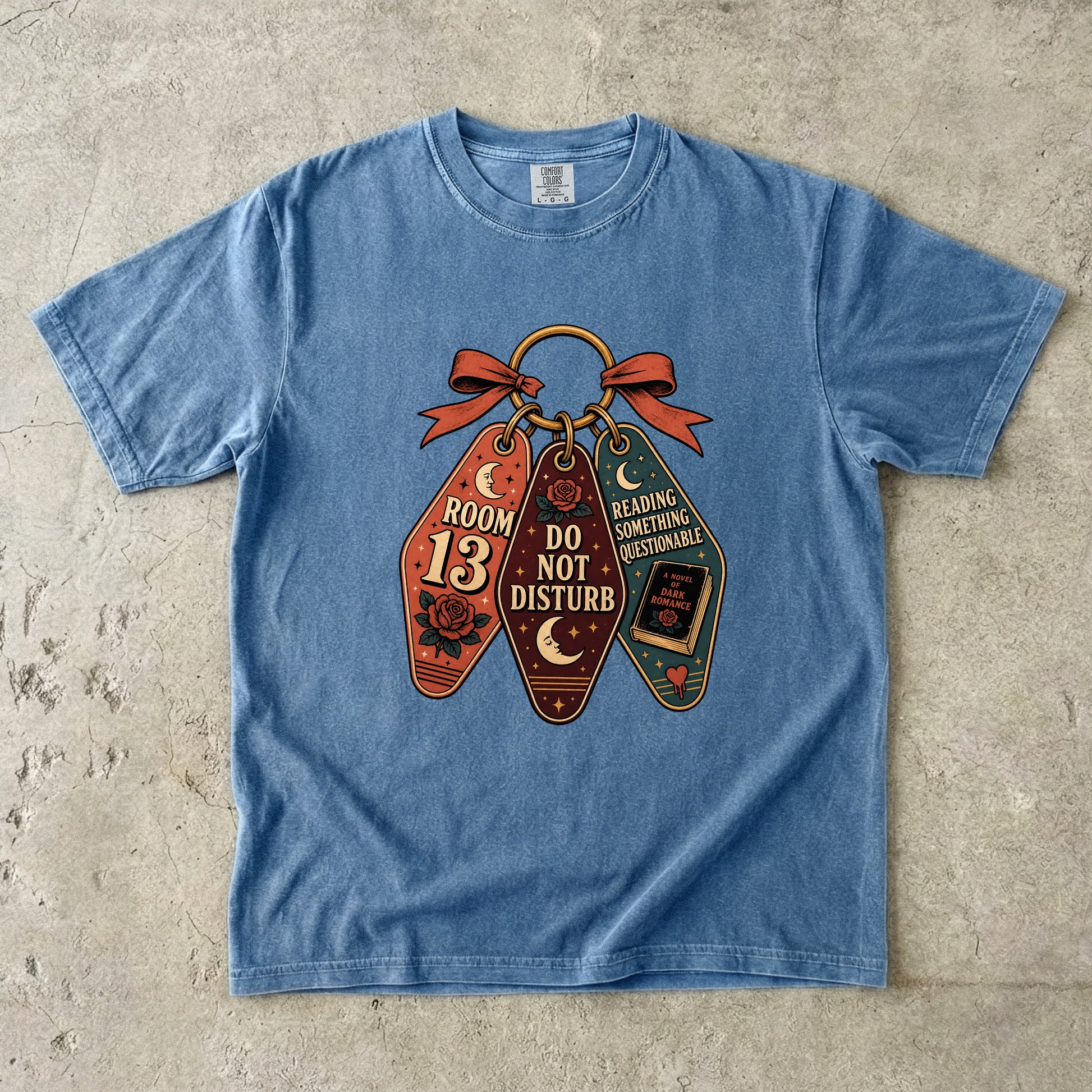 A light blue short-sleeve t-shirt laid flat on a textured concrete surface, featuring a graphic of three vintage-style library book tags tied with a red ribbon. The tags read 'ROOM 13 DO NOT DISTURB', 'READING SCHEDULE DETERMINABLE', and 'I HAVE A BOOK COMING FOR ME'.