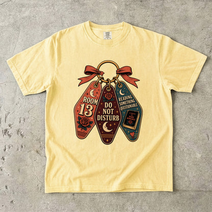 A pale yellow short-sleeve t-shirt laid flat on a textured gray concrete surface. The front of the shirt features a graphic of three hanging keychains tied with a red bow.