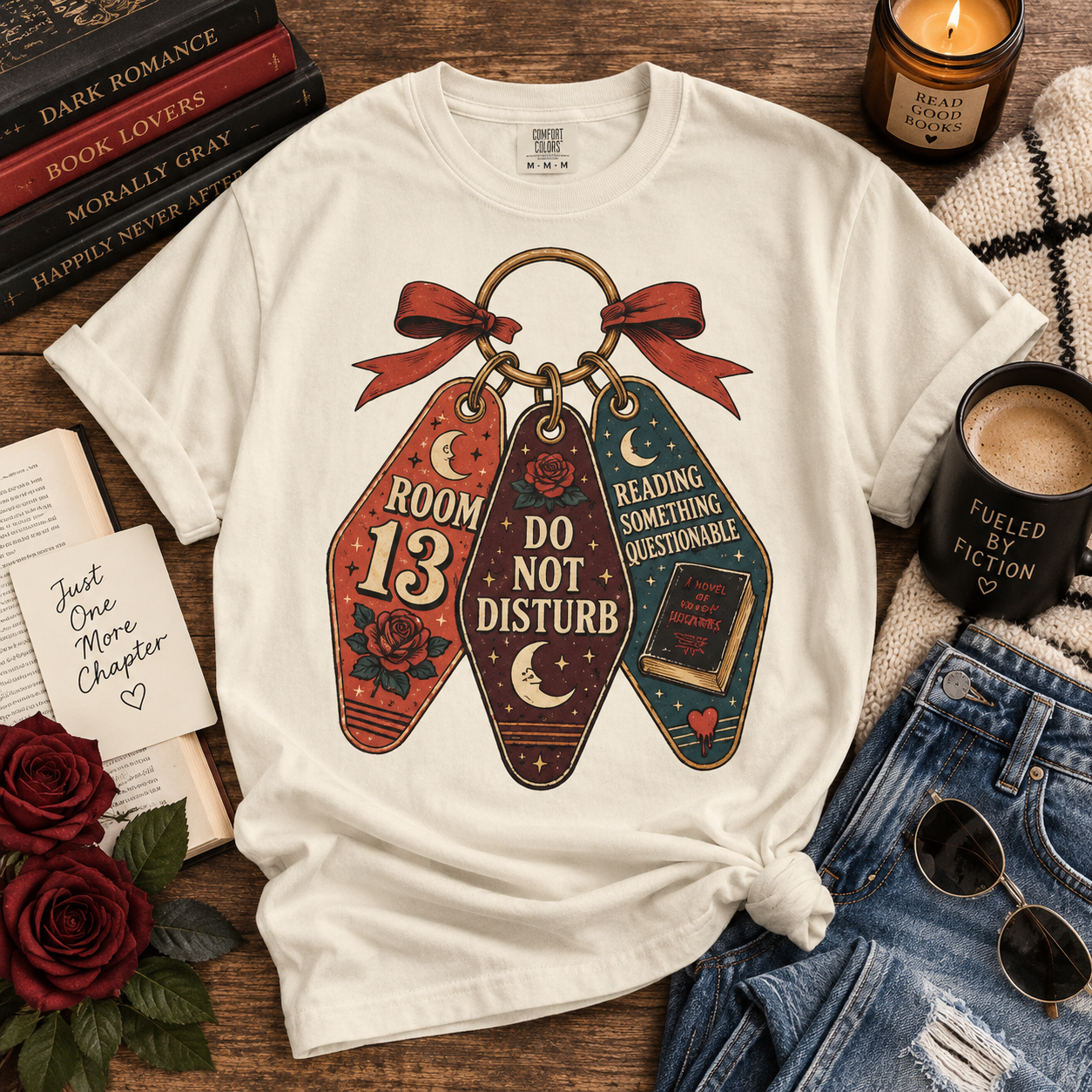 A white graphic T-shirt laid flat on a wooden surface, surrounded by styled props including books, a candle, a mug, sunglasses, and jeans. The shirt features a large central graphic of three hanging keychains tied with a red bow, with the words 'ROOM 13', 'DO NOT DISTURB', and 'READING SOMETHING ACCEPTABLE'.