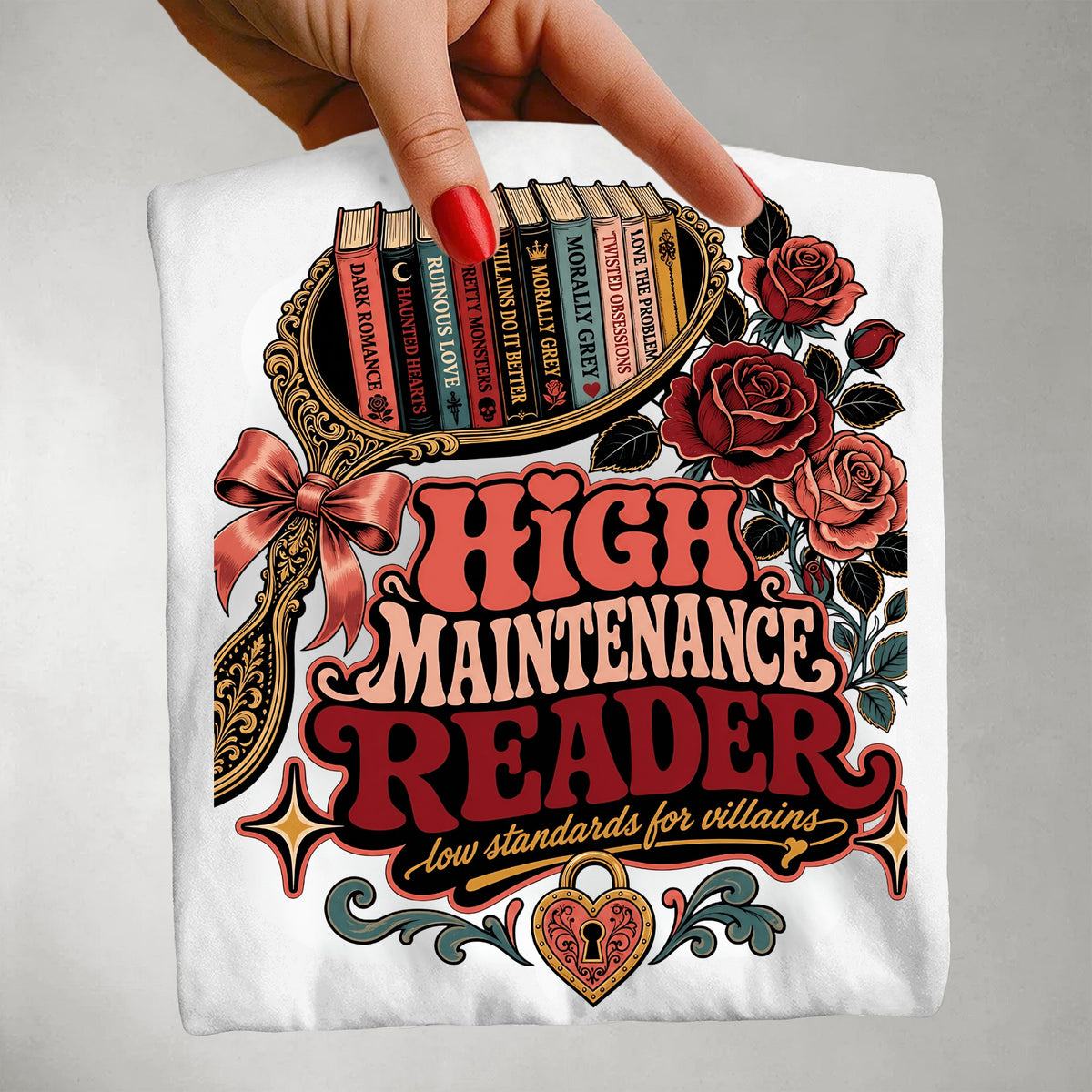 A hand with red nail polish holds up a white t-shirt featuring a detailed graphic print. The graphic reads 'HIGH MAINTENANCE READER' in red and pink retro lettering, with 'low standards for villains' underneath.