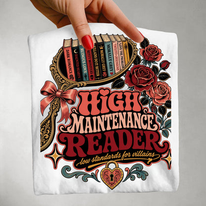 A hand with red nail polish holds up a white t-shirt featuring a detailed graphic print. The graphic reads 'HIGH MAINTENANCE READER' in red and pink retro lettering, with 'low standards for villains' underneath.