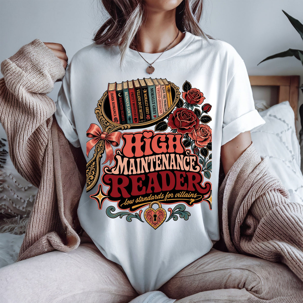 A person wearing a white graphic T-shirt with a large vintage-style book-themed print reading 'High Maintenance Reader' and 'Low standards for villains,' styled with roses, books, and decorative flourishes. A beige knit sweater is draped off the shoulders, and a bed with pillows is softly visible in the background.