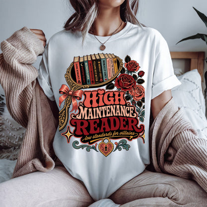 A person wearing a white graphic T-shirt with a large vintage-style book-themed print reading 'High Maintenance Reader' and 'Low standards for villains,' styled with roses, books, and decorative flourishes. A beige knit sweater is draped off the shoulders, and a bed with pillows is softly visible in the background.
