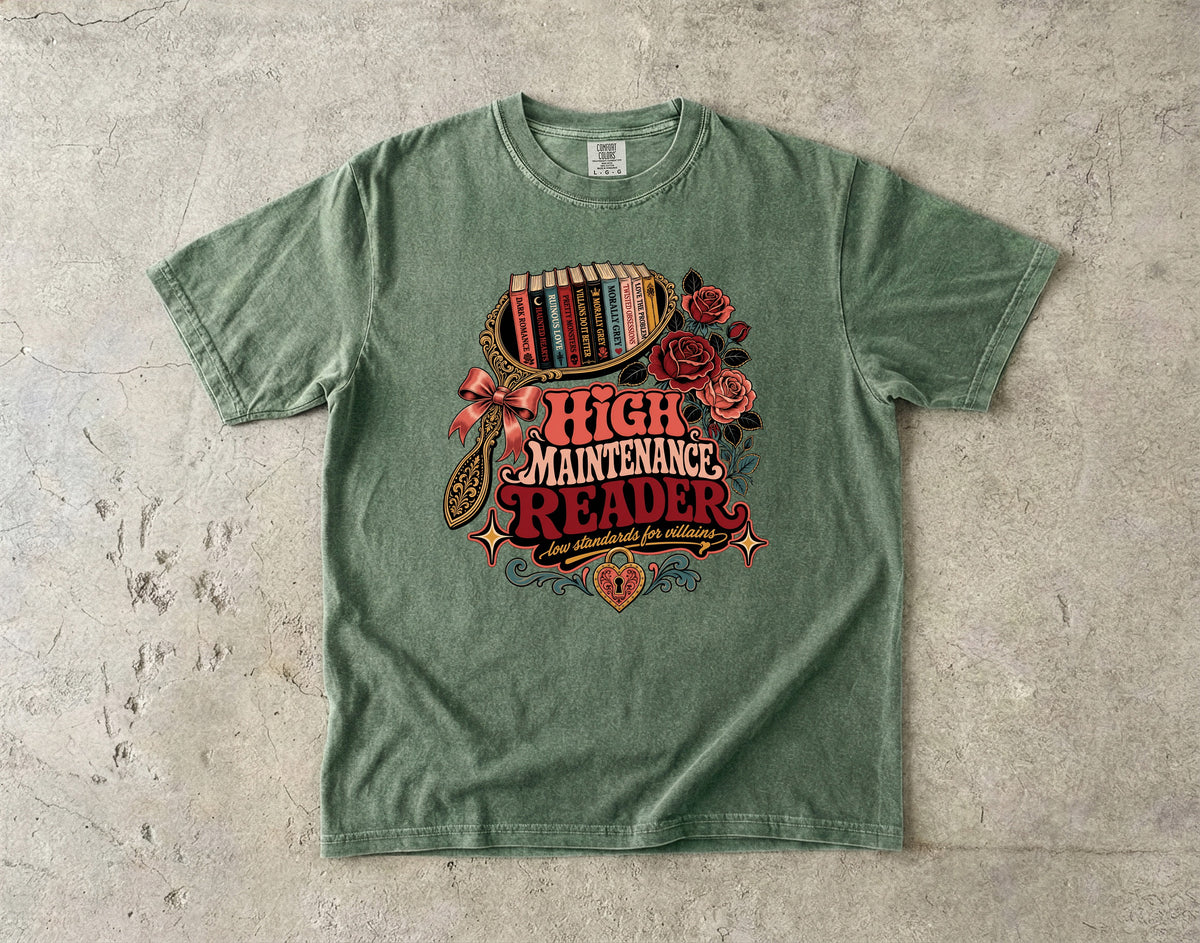 A sage green graphic t-shirt laid flat on a textured concrete surface. The shirt features a retro-style graphic with the words 'HIGH MAINTENANCE READER' in red and cream lettering, surrounded by illustrations of red roses, a bookshelf filled with books, and a bow.