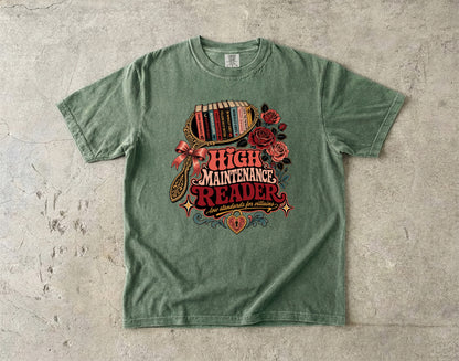 A sage green graphic t-shirt laid flat on a textured concrete surface. The shirt features a retro-style graphic with the words 'HIGH MAINTENANCE READER' in red and cream lettering, surrounded by illustrations of red roses, a bookshelf filled with books, and a bow.