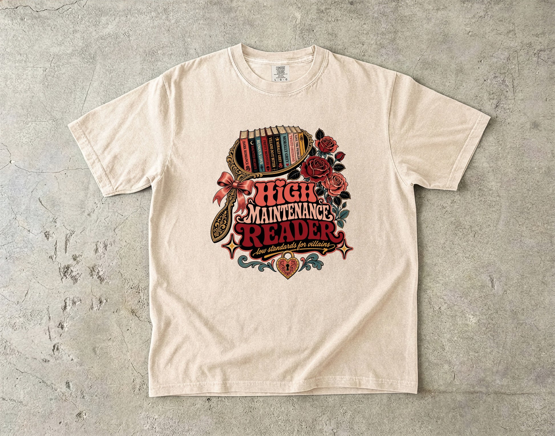 An off-white crewneck t-shirt with a vintage-style graphic reading 'HIGH MAINTENANCE READER' surrounded by roses, a bookshelf, and a magnifying glass, laid flat on a textured gray concrete surface.