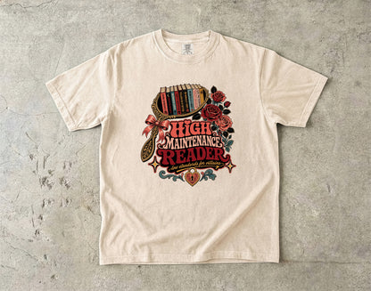An off-white crewneck t-shirt with a vintage-style graphic reading 'HIGH MAINTENANCE READER' surrounded by roses, a bookshelf, and a magnifying glass, laid flat on a textured gray concrete surface.