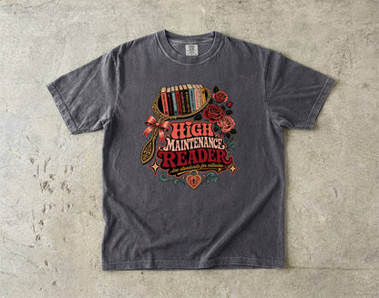 A dark gray heather t-shirt with a vintage-style 'High Maintenance Reader' graphic, laid flat on a textured concrete surface.