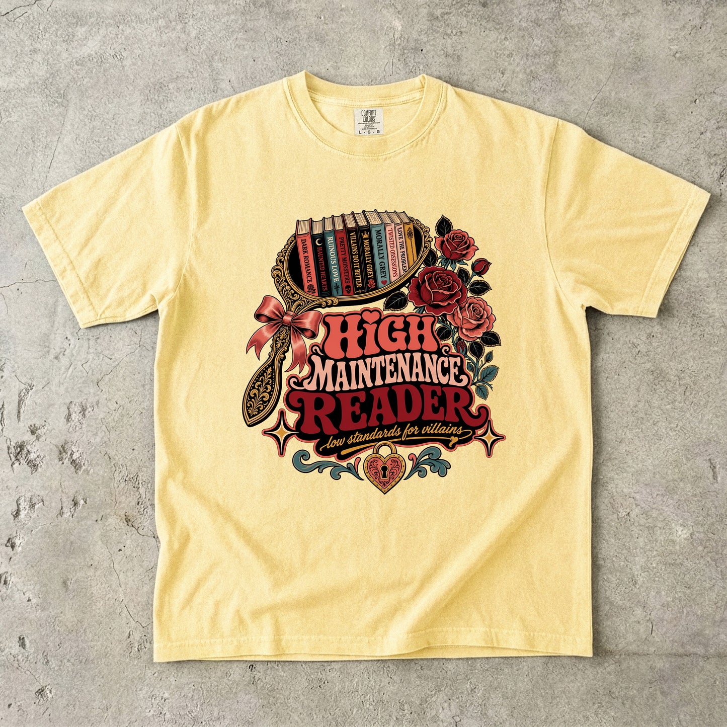 A pale yellow graphic t-shirt laid flat on a textured gray concrete background. The shirt features a large, detailed retro-style graphic on the chest depicting a bookshelf, roses, a bow, and the text 'HIGH MAINTENANCE READER' in red and white.