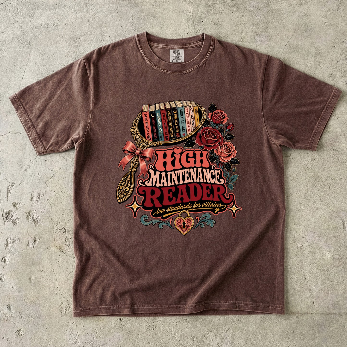 A brown short-sleeve graphic t-shirt laid flat on a textured gray concrete background. The shirt features a colorful retro-style illustration of a bookshelf filled with books, red roses, a bow, and the text 'HIGH MAINTENANCE READER' in red and orange lettering.
