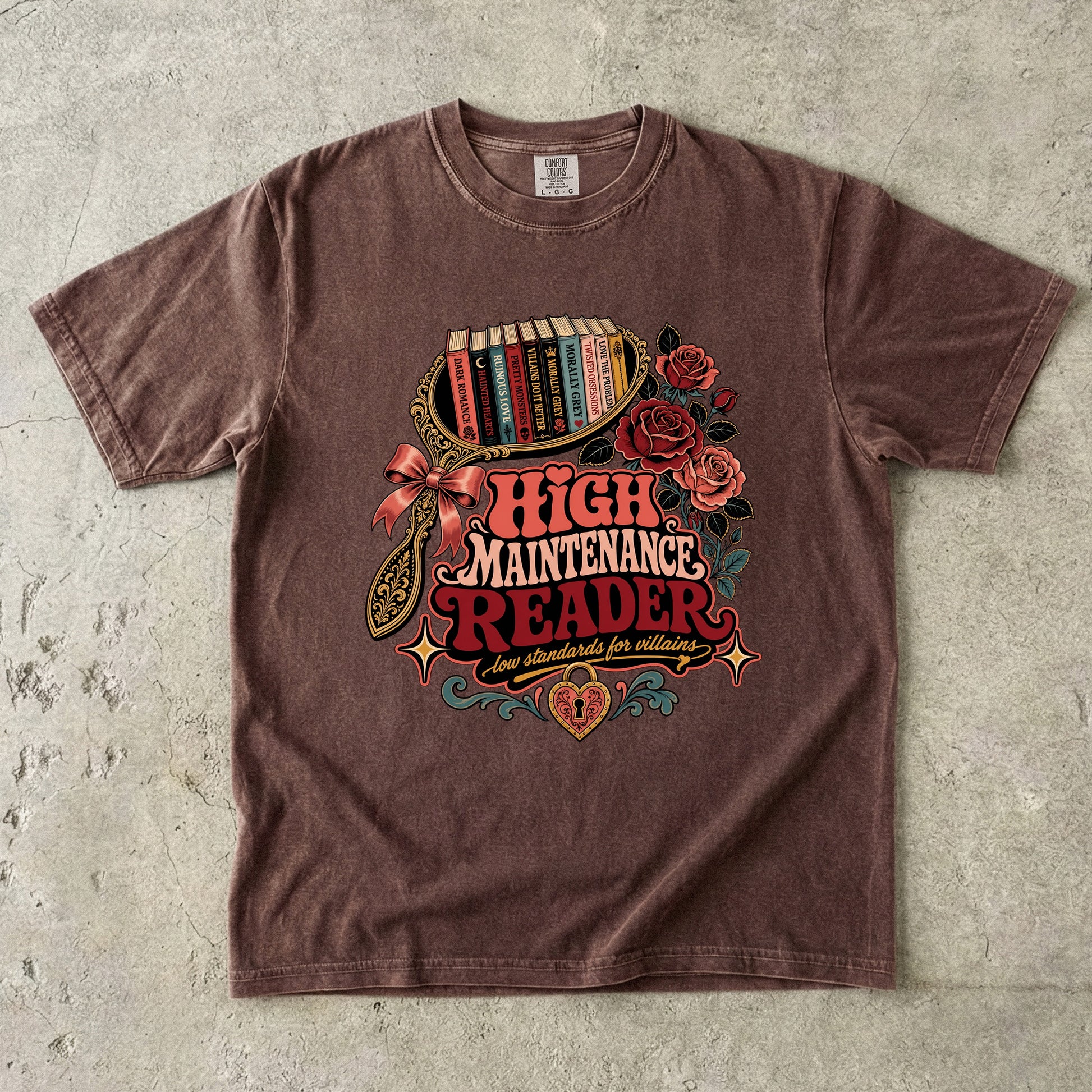 A brown short-sleeve graphic t-shirt laid flat on a textured gray concrete background. The shirt features a colorful retro-style illustration of a bookshelf filled with books, red roses, a bow, and the text 'HIGH MAINTENANCE READER' in red and orange lettering.