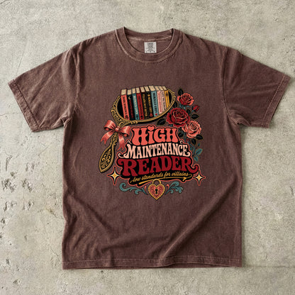 A brown short-sleeve graphic t-shirt laid flat on a textured gray concrete background. The shirt features a colorful retro-style illustration of a bookshelf filled with books, red roses, a bow, and the text 'HIGH MAINTENANCE READER' in red and orange lettering.