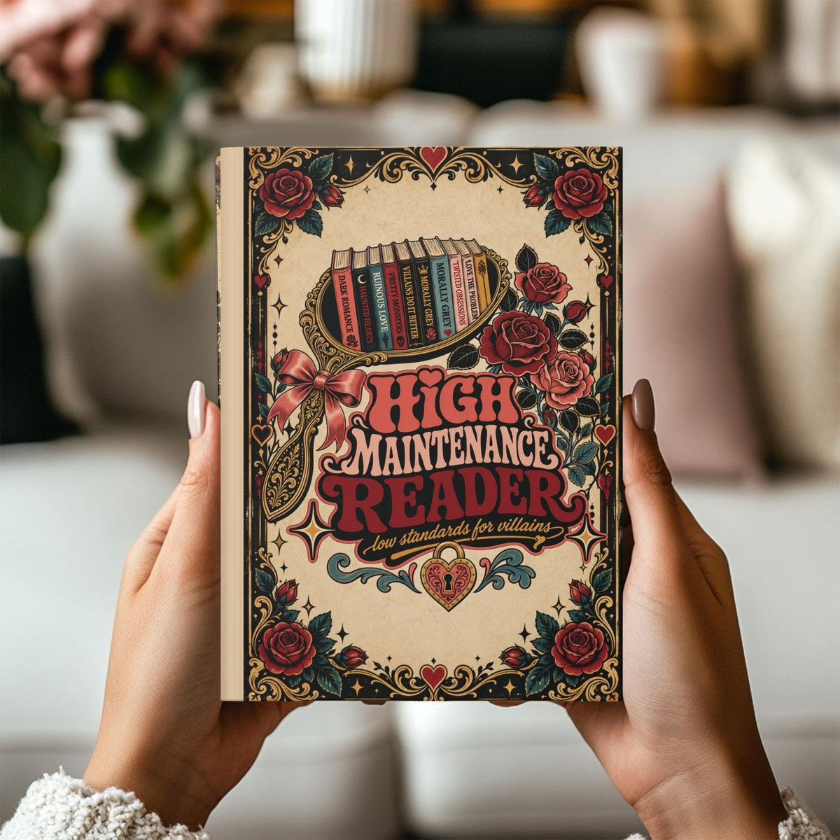 A person holds a hardcover book titled "High Maintenance Reader" with a vintage-style illustrated cover featuring roses, hearts, and a magnifying glass over stacked books. The background is a softly blurred indoor setting with neutral tones.