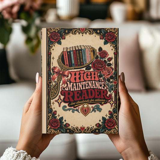 A person holds a hardcover book titled "High Maintenance Reader" with a vintage-style illustrated cover featuring roses, hearts, and a magnifying glass over stacked books. The background is a softly blurred indoor setting with neutral tones.