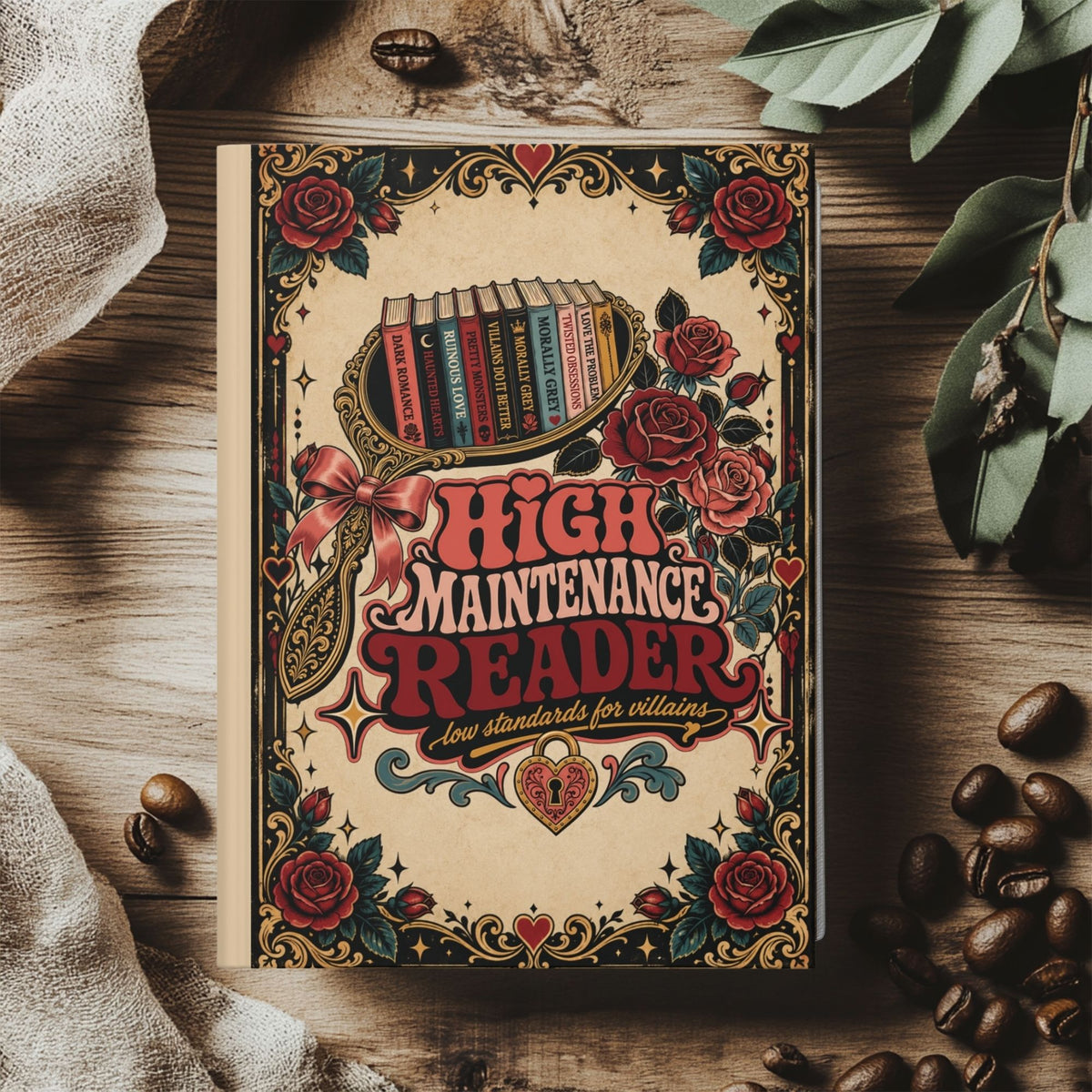 A decorative book cover or notebook titled "High Maintenance Reader" lies on a rustic wooden surface, surrounded by coffee beans, a cloth, and green leaves. The cover features a vintage tattoo-style design with red roses, ornate borders, and a small illustration of a stack of books.