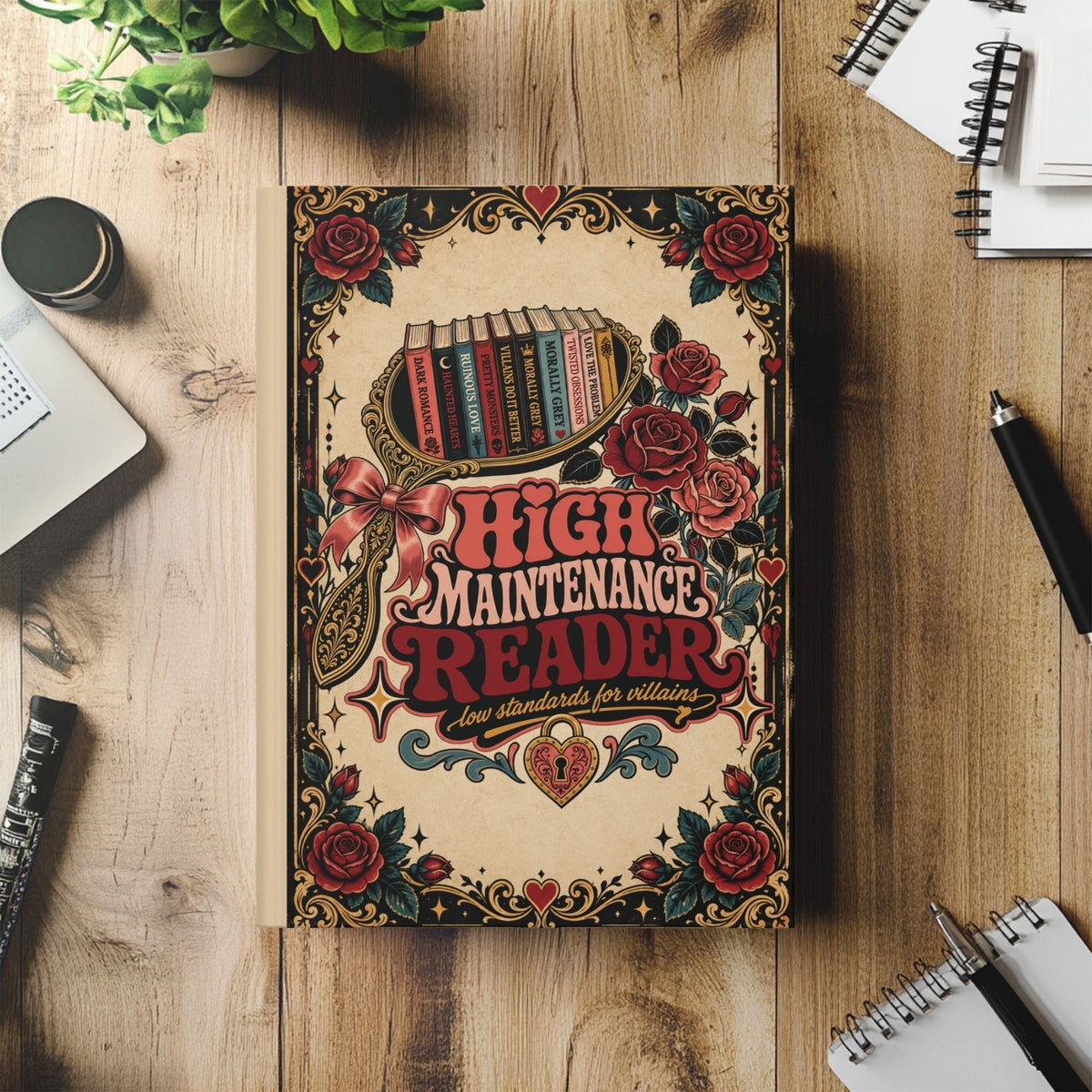 A decorative book titled "High Maintenance Reader" lies on a wooden desk surrounded by notebooks, a pen, a plant, and a laptop. The cover features a vintage tattoo-style design with red roses, hearts, and a bookshelf illustration.