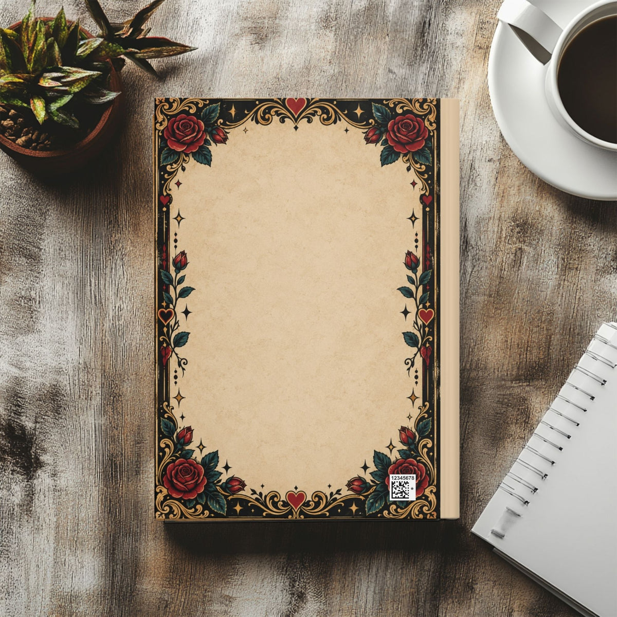 A decorative notebook with a vintage-style floral border featuring red roses and gold accents on a beige page, styled on a wooden tabletop with a plant, coffee cup, and spiral notebook nearby.