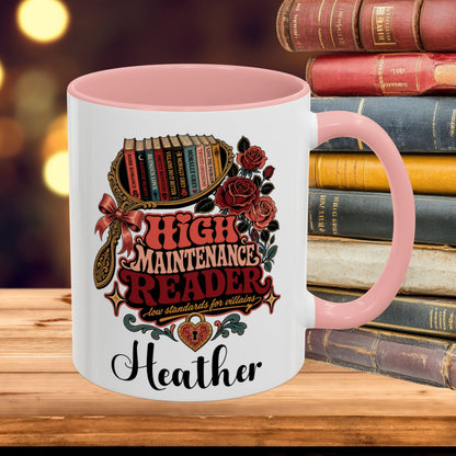 A white ceramic mug with a pink interior and handle, featuring a decorative graphic of books, roses, and the text 'HIGH MAINTENANCE READER' and the name 'Heather'. The mug is placed on a wooden surface with a stack of vintage books in the background.