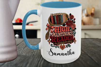 A white ceramic mug with a blue handle and interior, featuring a vintage-style book-themed graphic that reads 'High Maintenance Reader' and the name 'Samantha' printed below, shown on a wooden tabletop with a blurred kitchen background.