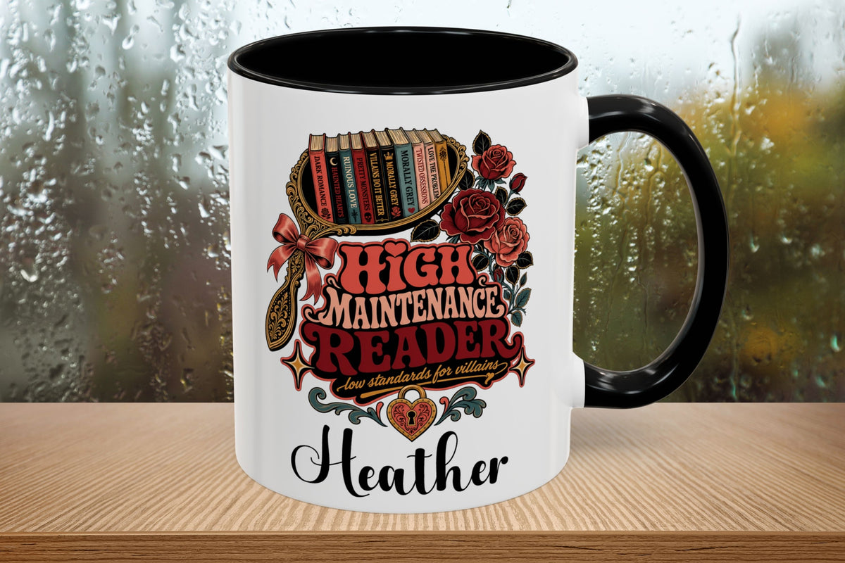 A white ceramic mug with a black handle and black interior, featuring a colorful graphic of a bookshelf, roses, and the text 'HIGH MAINTENANCE READER' with 'Heather' printed below, set on a wooden surface in front of a rainy window.