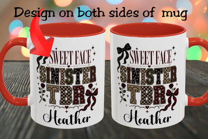 Two views of a white ceramic mug with a red handle and red interior, shown on a dark wooden surface with colorful blurred lights in the background. Text at the top says 'Design on both sides of mug' with a red arrow pointing to the left mug, which features the printed design: 'SWEET FACE SINISTER TDR Heather'.