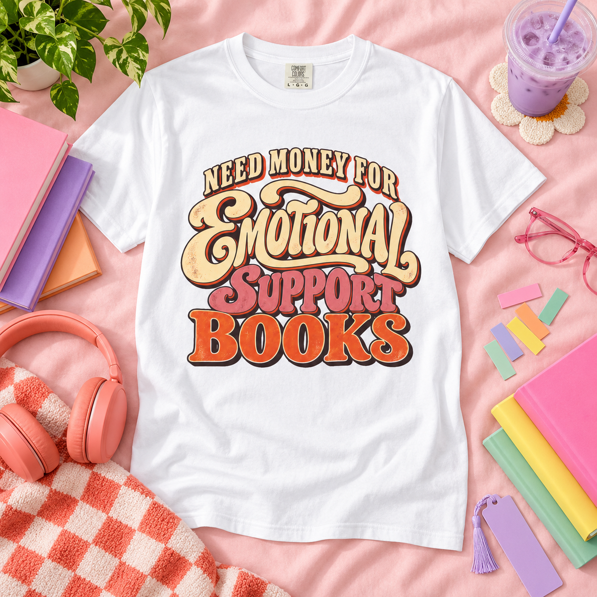 A white graphic T-shirt laid flat on a pink fabric background, surrounded by books, headphones, glasses, a drink, and a plant. The shirt features a large retro-style text graphic reading “Need Money for Emotional Support Books.