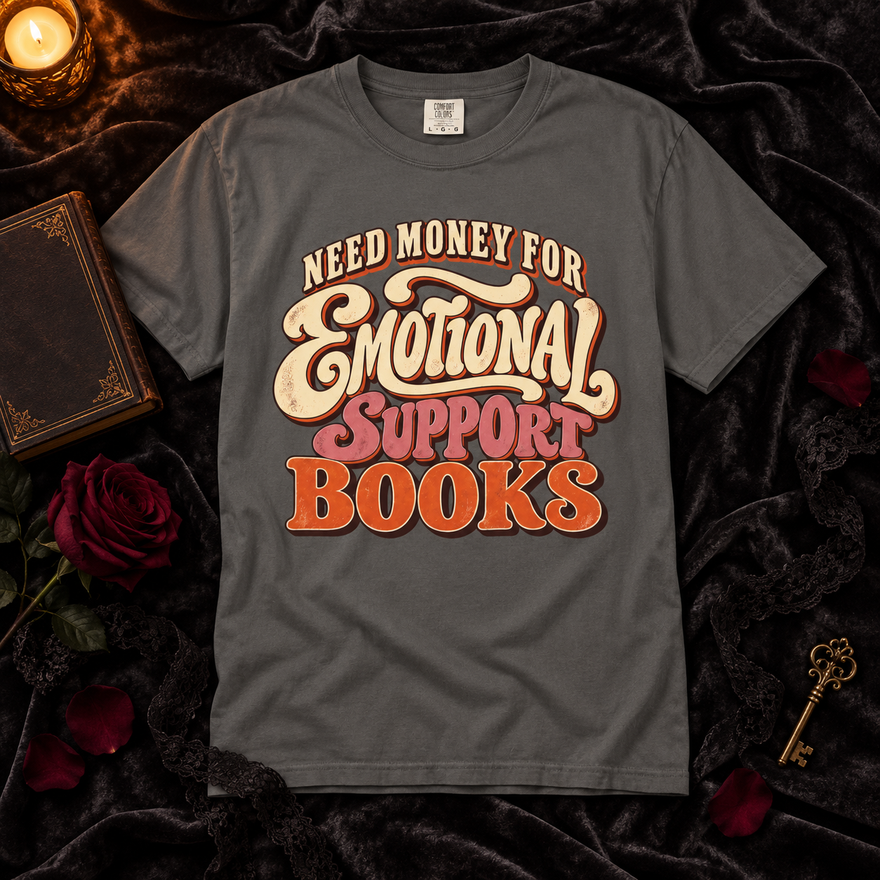 A dark gray graphic t-shirt laid flat on a black velvet background with decorative props including a candle, old book, roses, and a key. The shirt features a retro-style text graphic reading 'NEED MONEY FOR Emotional SUPPORT BOOKS' in cream, pink, and orange lettering.