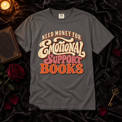A dark gray graphic t-shirt laid flat on a black velvet background with decorative props including a candle, old book, roses, and a key. The shirt features a retro-style text graphic reading 'NEED MONEY FOR Emotional SUPPORT BOOKS' in cream, pink, and orange lettering.